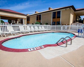 A swimming pool with a white fence, lounge chairs, and a building with a beige facade and a red roof.
