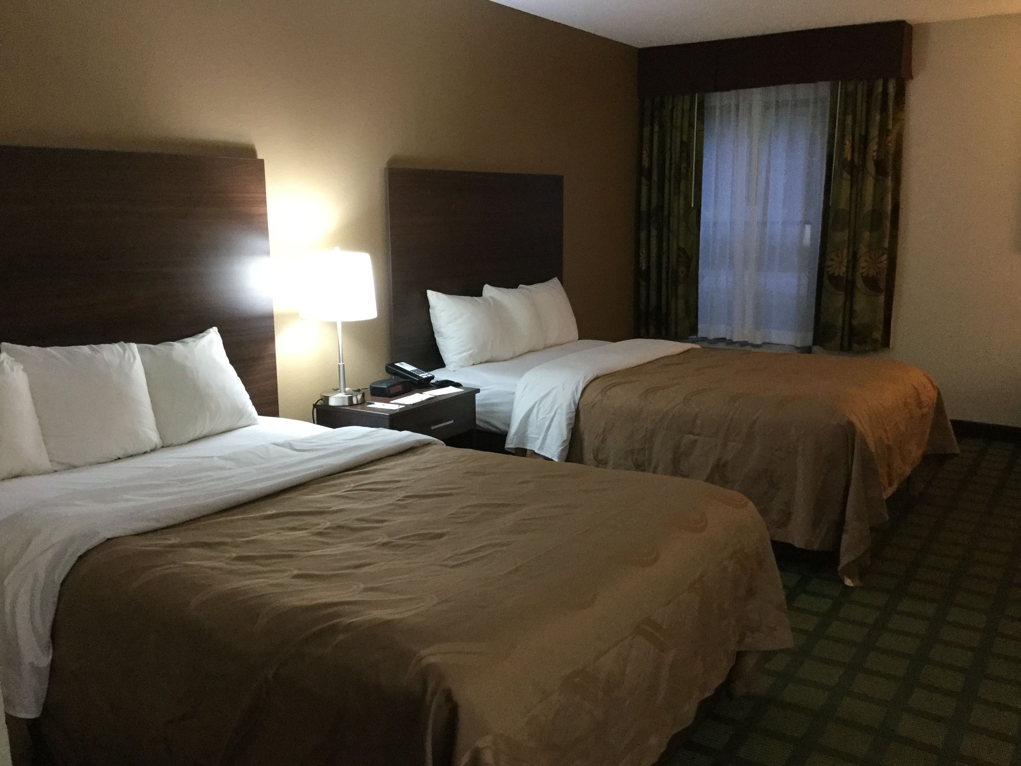 Habitación de hotel con dos camas, una mesita de noche con lámpara y una ventana con cortinas.