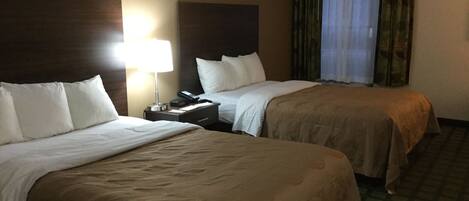Habitación de hotel con dos camas, una mesita de noche con lámpara y una ventana con cortinas.