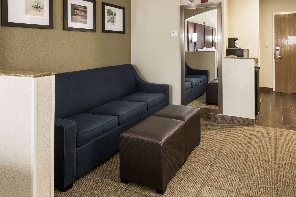 A hotel room with a dark blue sofa, a brown ottoman, and a small white table. There are framed pictures on the wall and a mirror above the table.