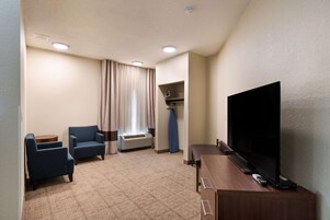 Quarto de hotel com televisão de tela plana, duas poltronas, uma mesinha, sofá, janela com cortinas e prateleira embutida.