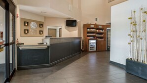 Uma área de recepção de hotel, com balcão de atendimento, televisão na parede, refrigerador e uma planta decorativa.