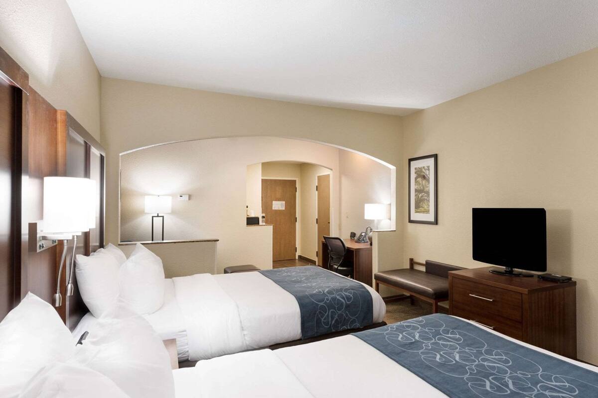 A hotel room with two beds, a desk, a chair, a television, and a bathroom visible through an archway.