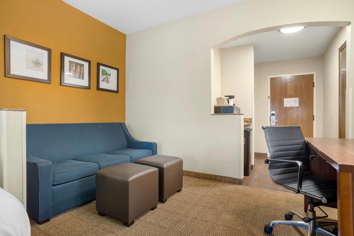 A hotel room with a blue sofa, two ottomans, a wooden desk, a black office chair, and framed pictures on the wall.