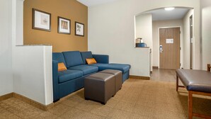 Uma sala de estar com um sofá azul, um banquinho marrom e uma mesa de jantar com cadeiras. Há quadros nas paredes e uma porta ao fundo.