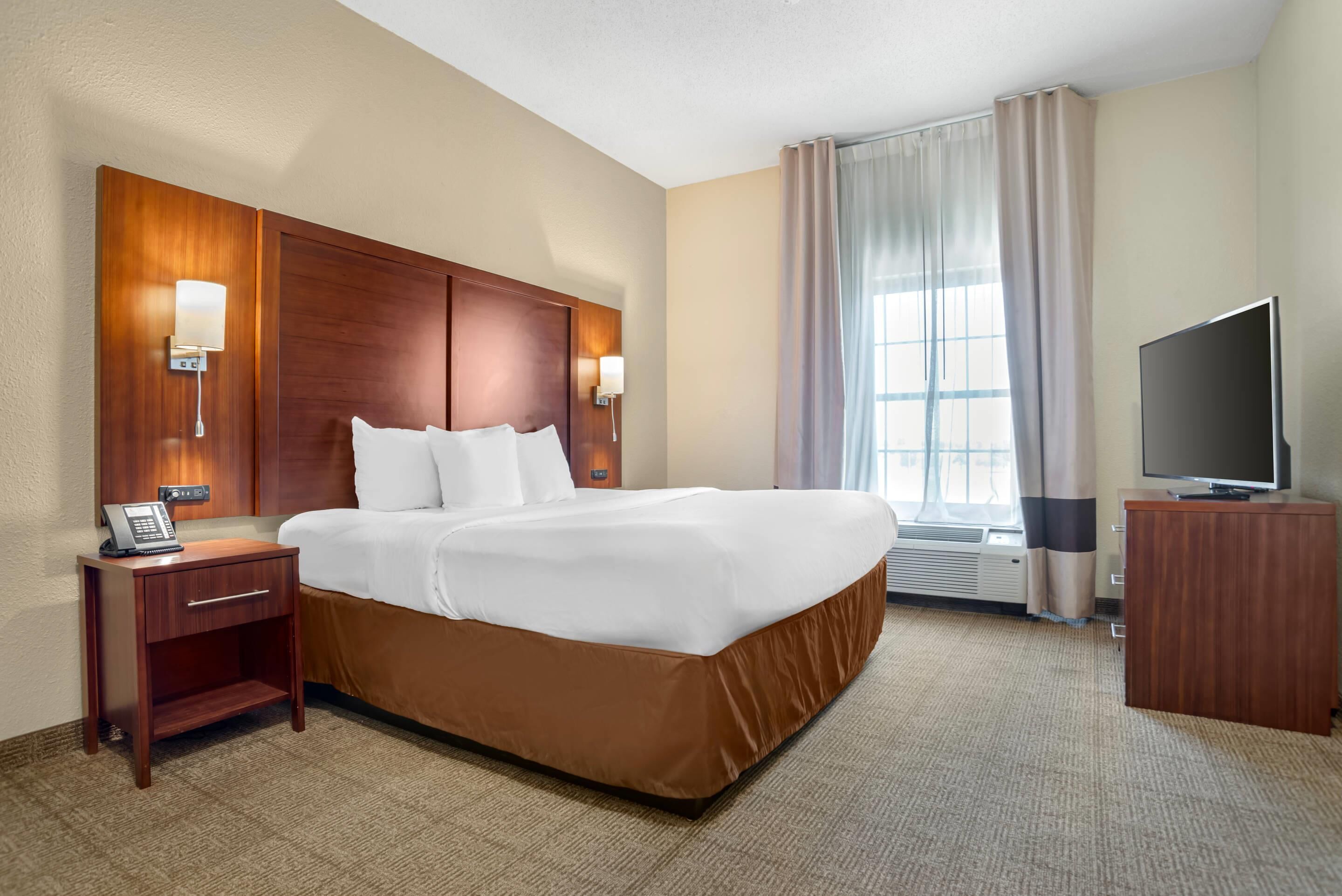 Una habitación de hotel con una cama grande, cabecera de madera, lámparas de noche, un escritorio con teléfono, televisión y una ventana con cortinas.