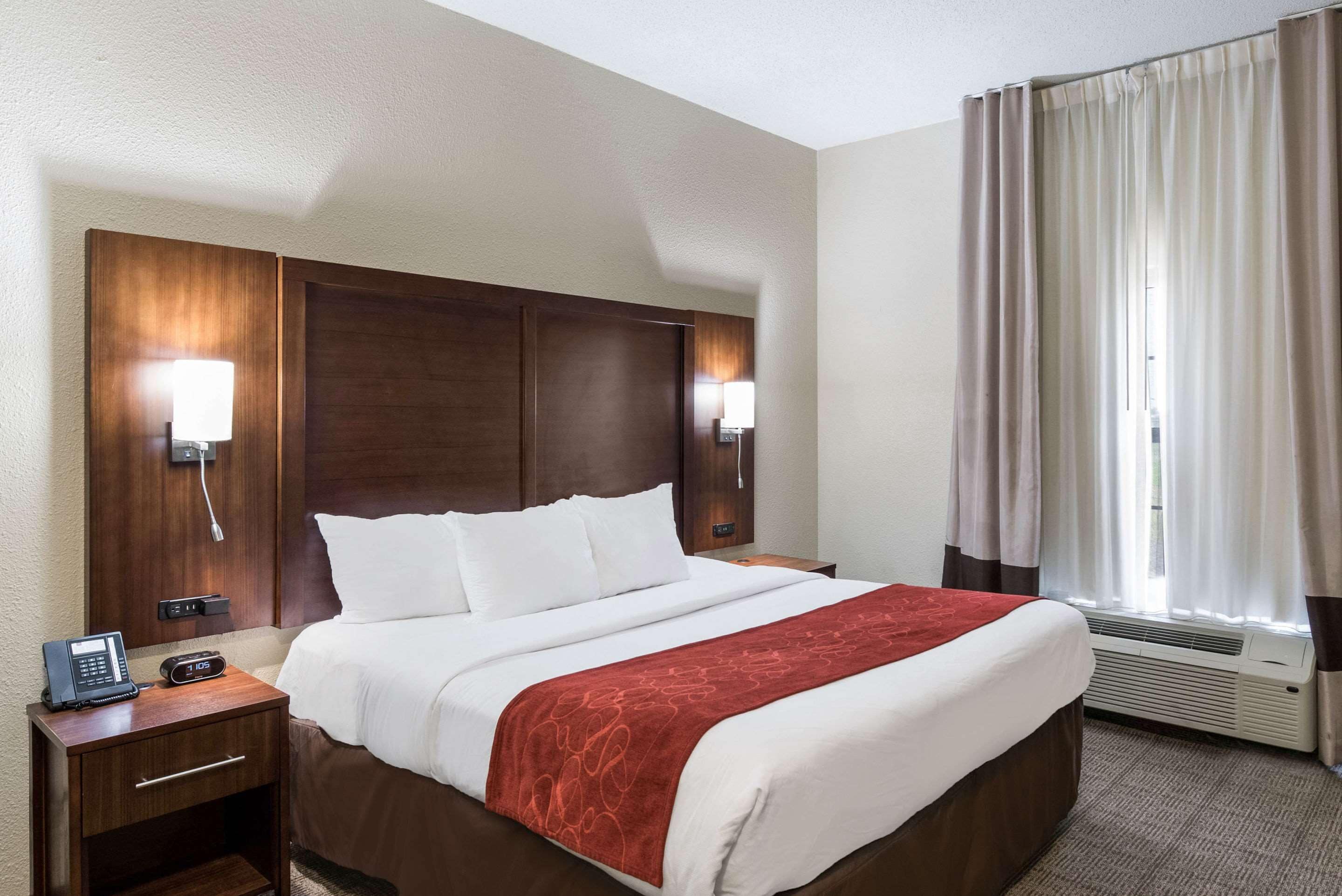 Ein Hotelzimmer mit einem großen Bett, einem Holzkopfstück, Nachttischlampen, einem Telefon und einem Fenster mit Vorhängen.
