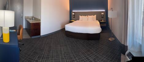 Una habitación de hotel con cama, un escritorio con lámpara, un baño pequeño y una ventana.