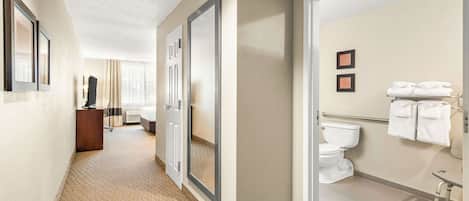 A hotel hallway with a carpeted floor, a wooden desk, a television, and a bathroom with a toilet, sink, and towels.