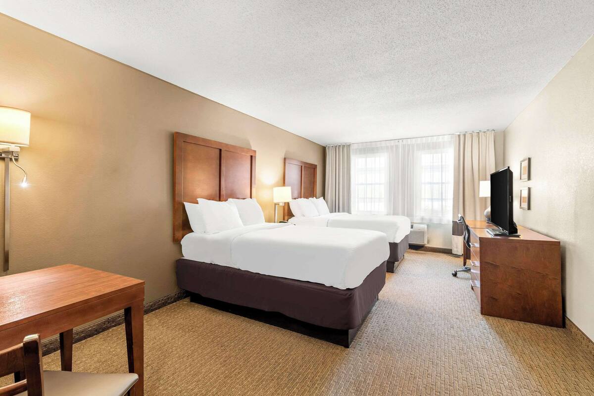 A hotel room with two beds, a desk, a chair, a TV, and a lamp.