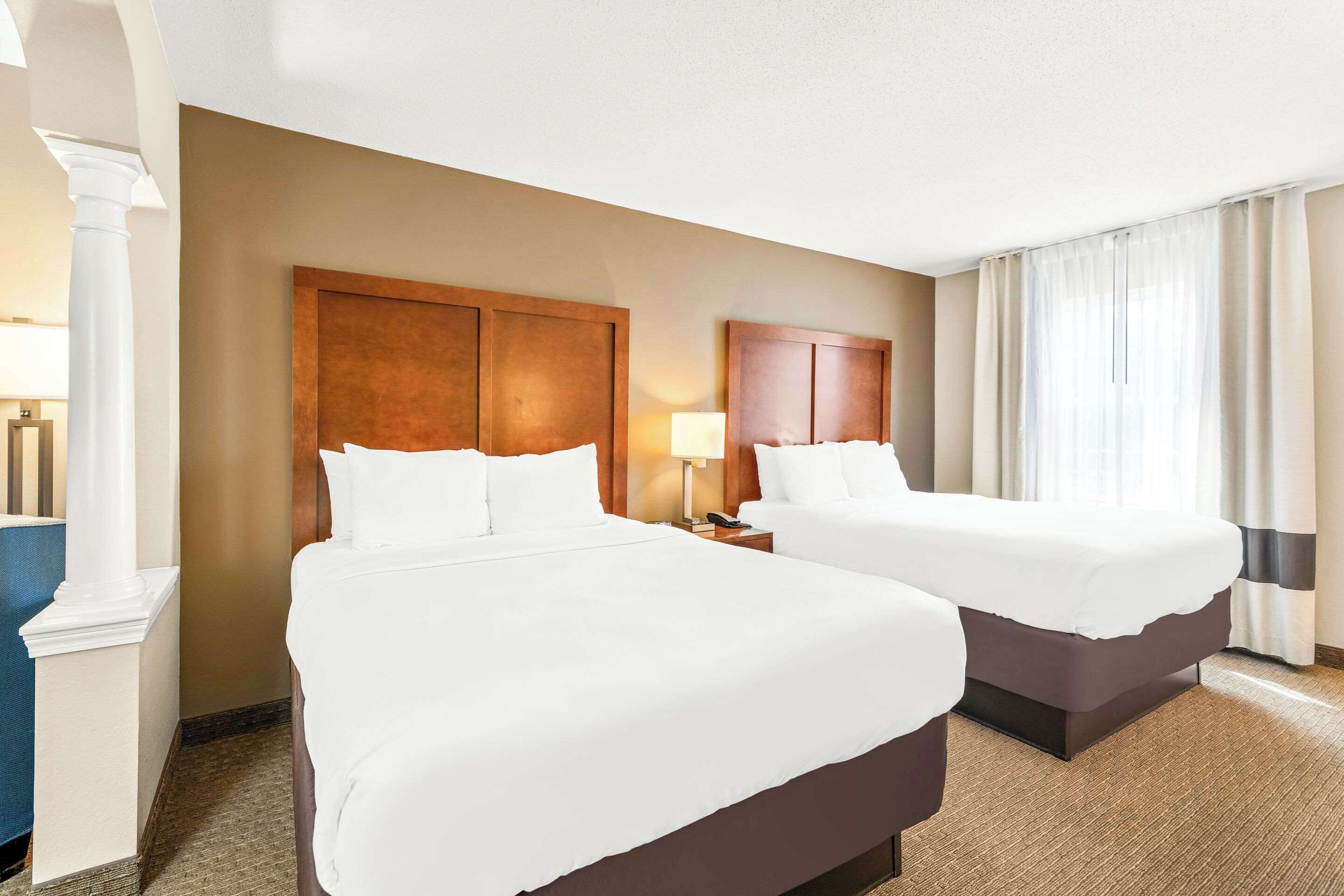 A hotel room with two beds, a large wooden headboard, a nightstand with a lamp, and a large window with curtains.