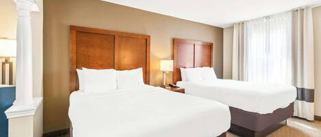 A hotel room with two beds, a large wooden headboard, a nightstand with a lamp, and a large window with curtains.
