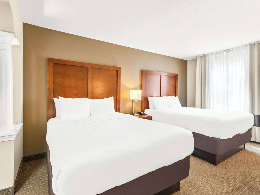 A hotel room with two beds, a large wooden headboard, a nightstand with a lamp, and a large window with curtains.