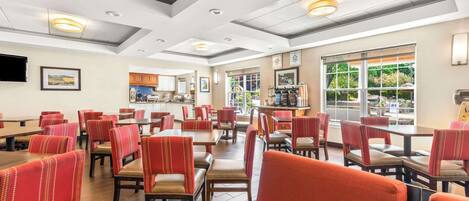 A spacious dining area with multiple tables and chairs, a kitchen in the background, and large windows providing natural light.