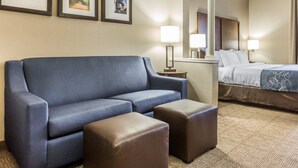 A hotel room with a blue sofa, two brown ottomans, a bed with white linens, and three framed pictures on the wall. - Comfort Suites at Isle Of Palms Connector (Mount Pleasant)