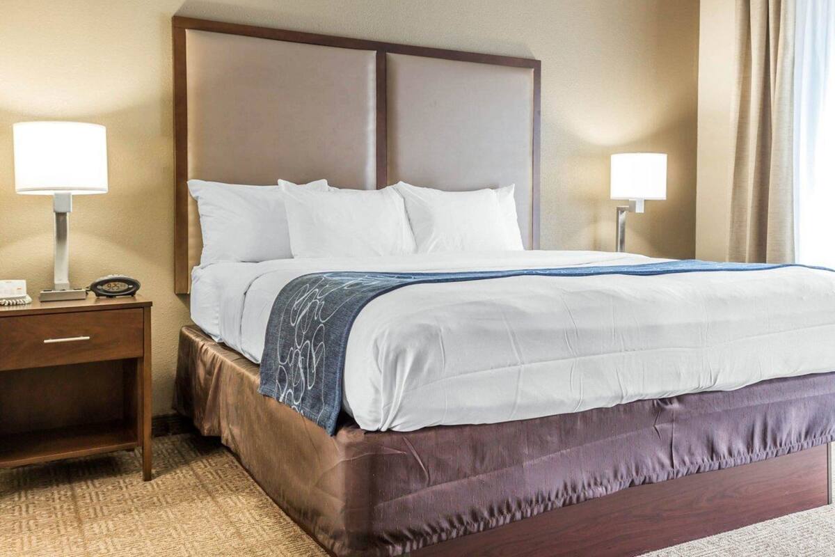 A hotel room with a large bed, two bedside tables with lamps, a wooden headboard, and a view of the outdoors through a window with curtains.