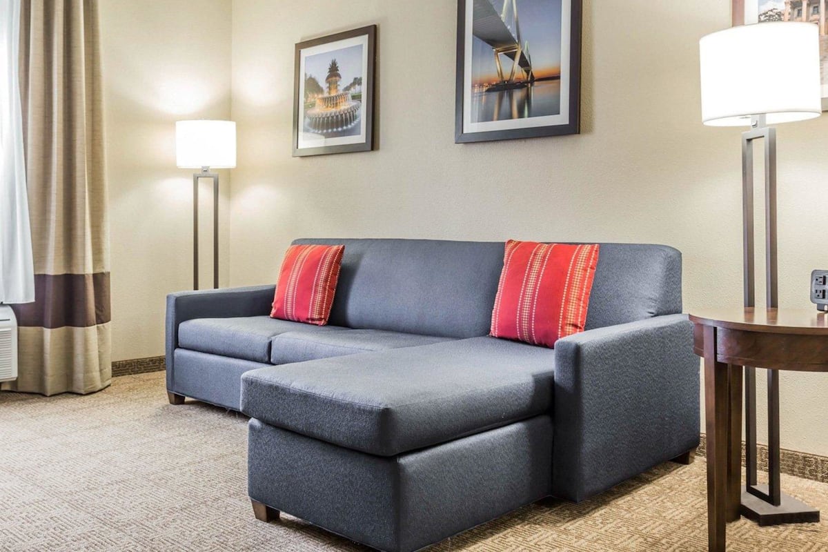 A modern hotel room with a grey sectional sofa, two red pillows, a standing floor lamp, and two framed pictures on the wall.