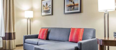 A modern hotel room with a grey sectional sofa, two red pillows, a standing floor lamp, and two framed pictures on the wall.