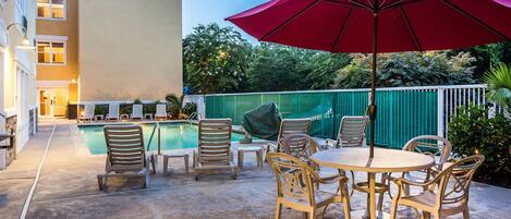 A poolside area with lounge chairs and a table, surrounded by a green fence and trees.