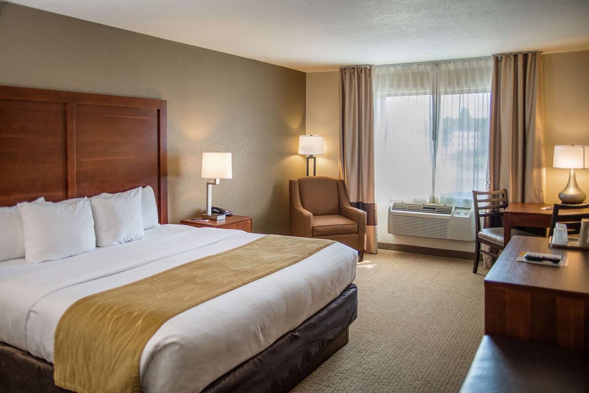 A hotel room with a large bed, a desk, a chair, and an air conditioning unit.