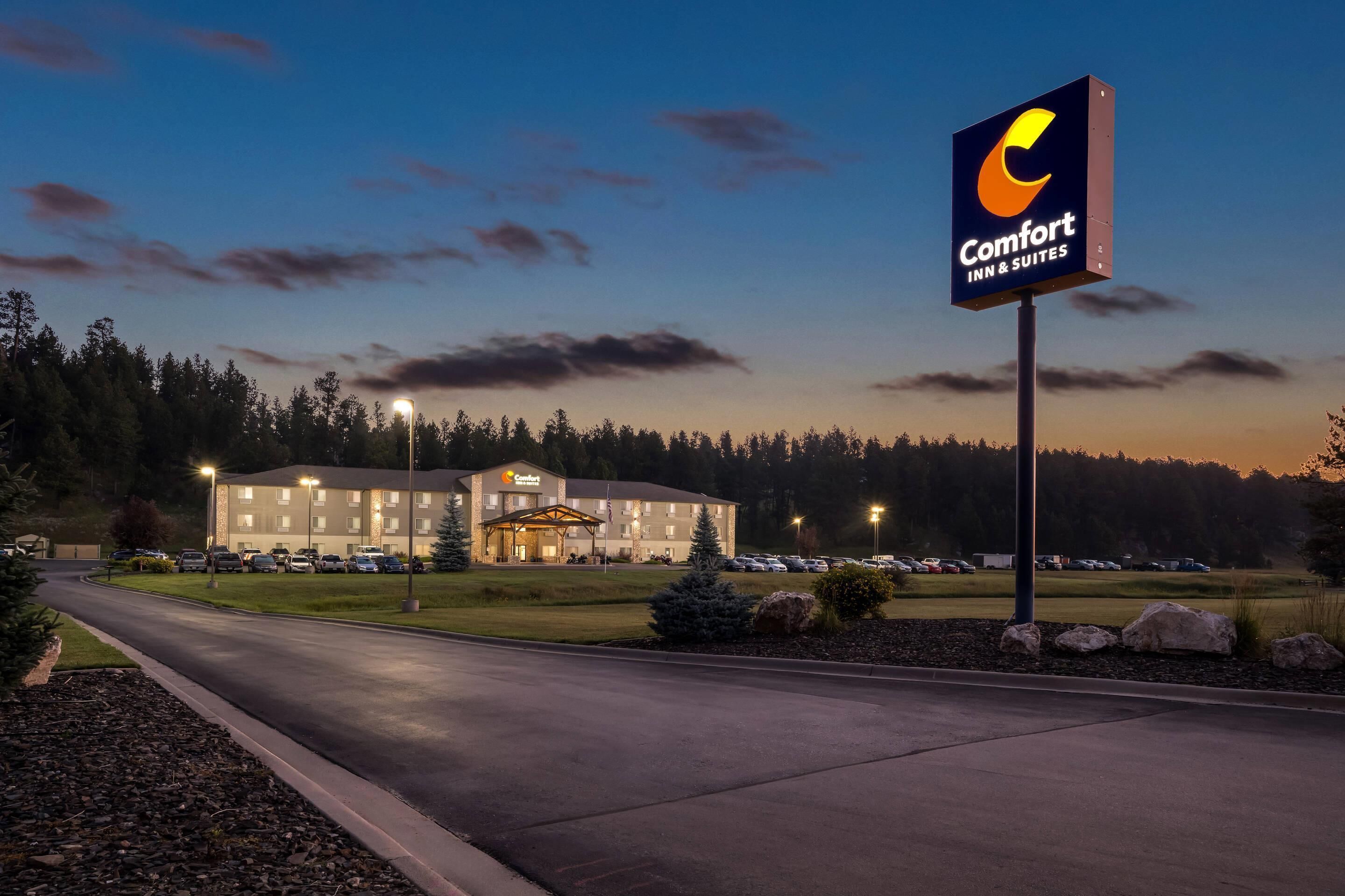 A Comfort Inn & Suites hotel with a well-lit entrance, a parking lot, and a clear view of the surrounding trees and sky.