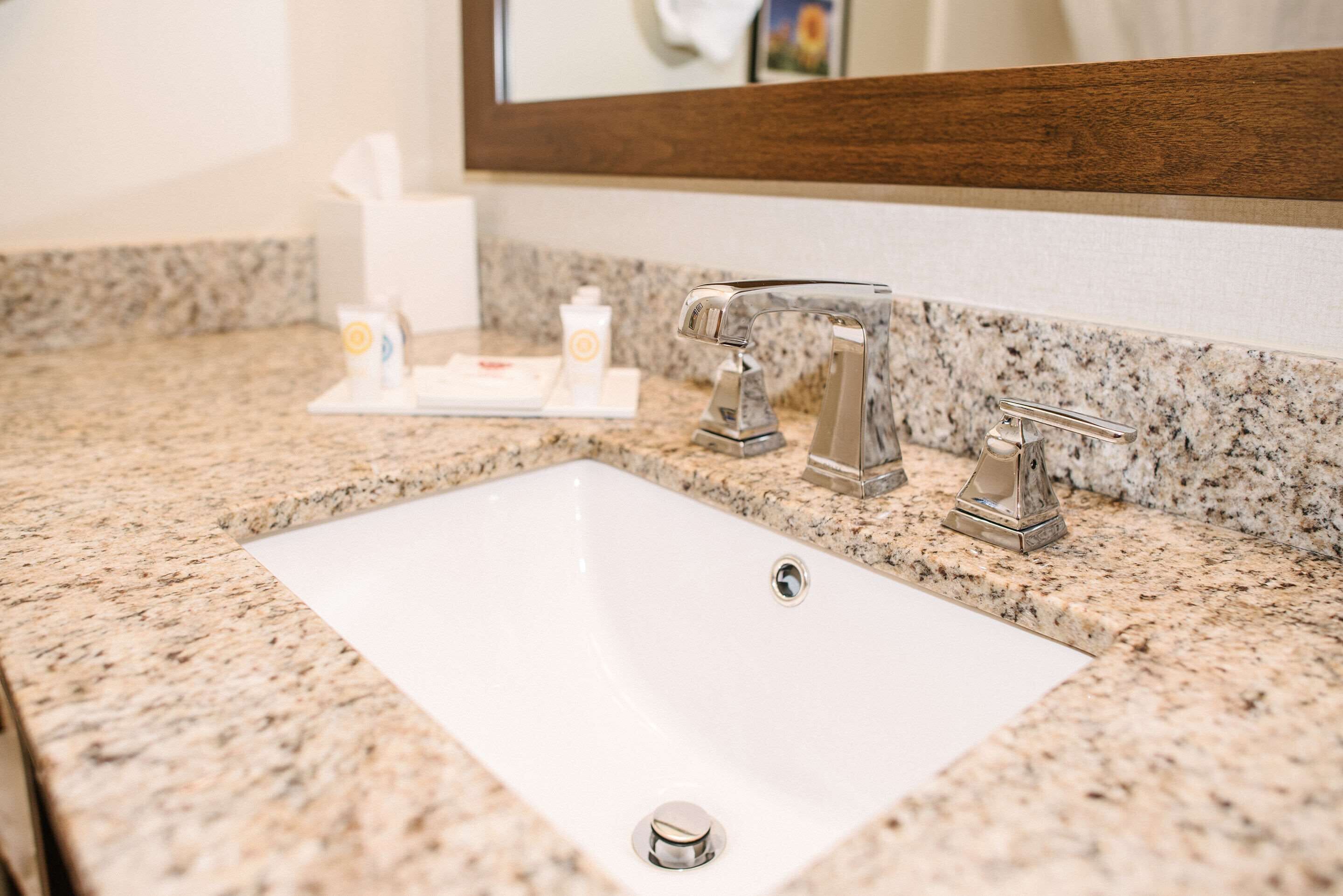 A bathroom sink with a granite countertop, a mirror, and a towel dispenser.