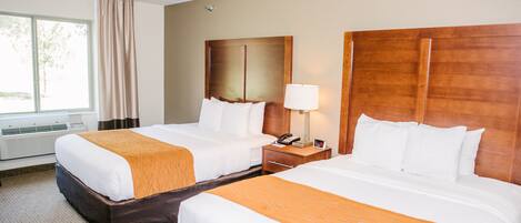 A hotel room with two beds, a window with curtains, a bedside table with a lamp, and a wall-mounted light fixture.