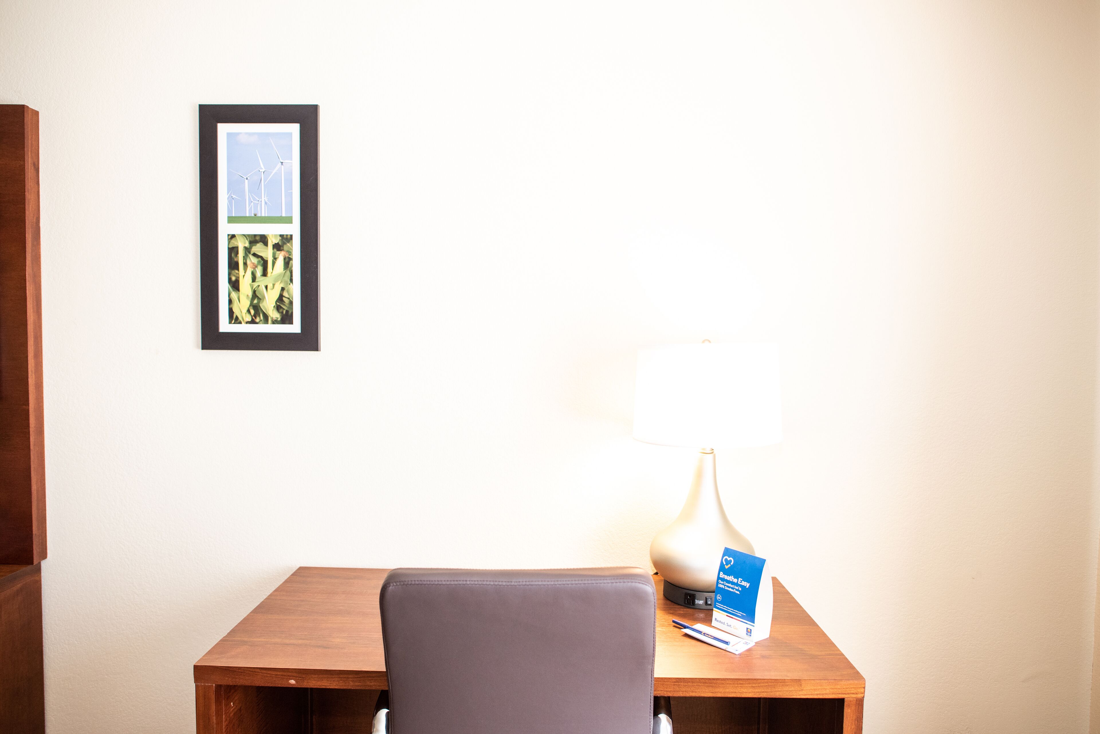 Quarto de hotel com escrivaninha de madeira, um quadro com turbinas e uma luminária com um folheto.