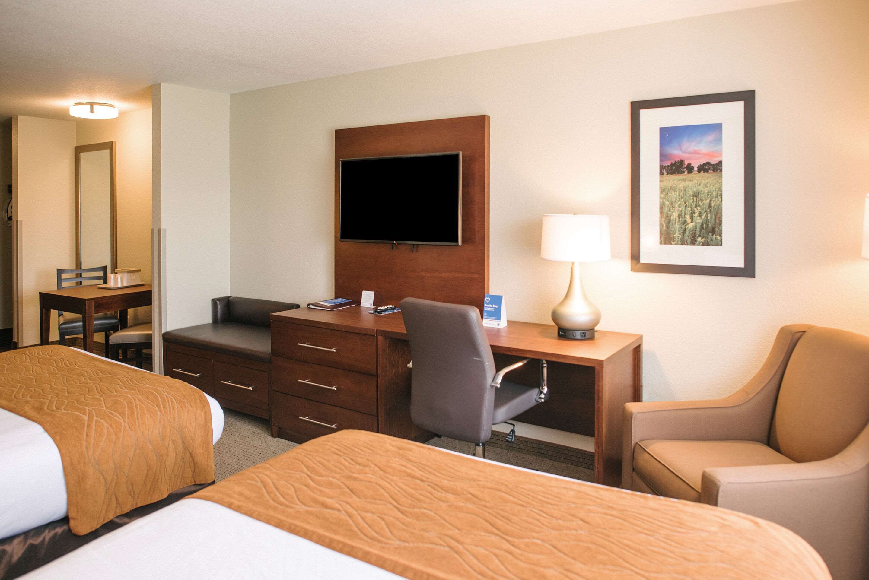 A hotel room with two beds, a desk with a flat-screen TV, a chair, a lamp, and a framed picture on the wall.