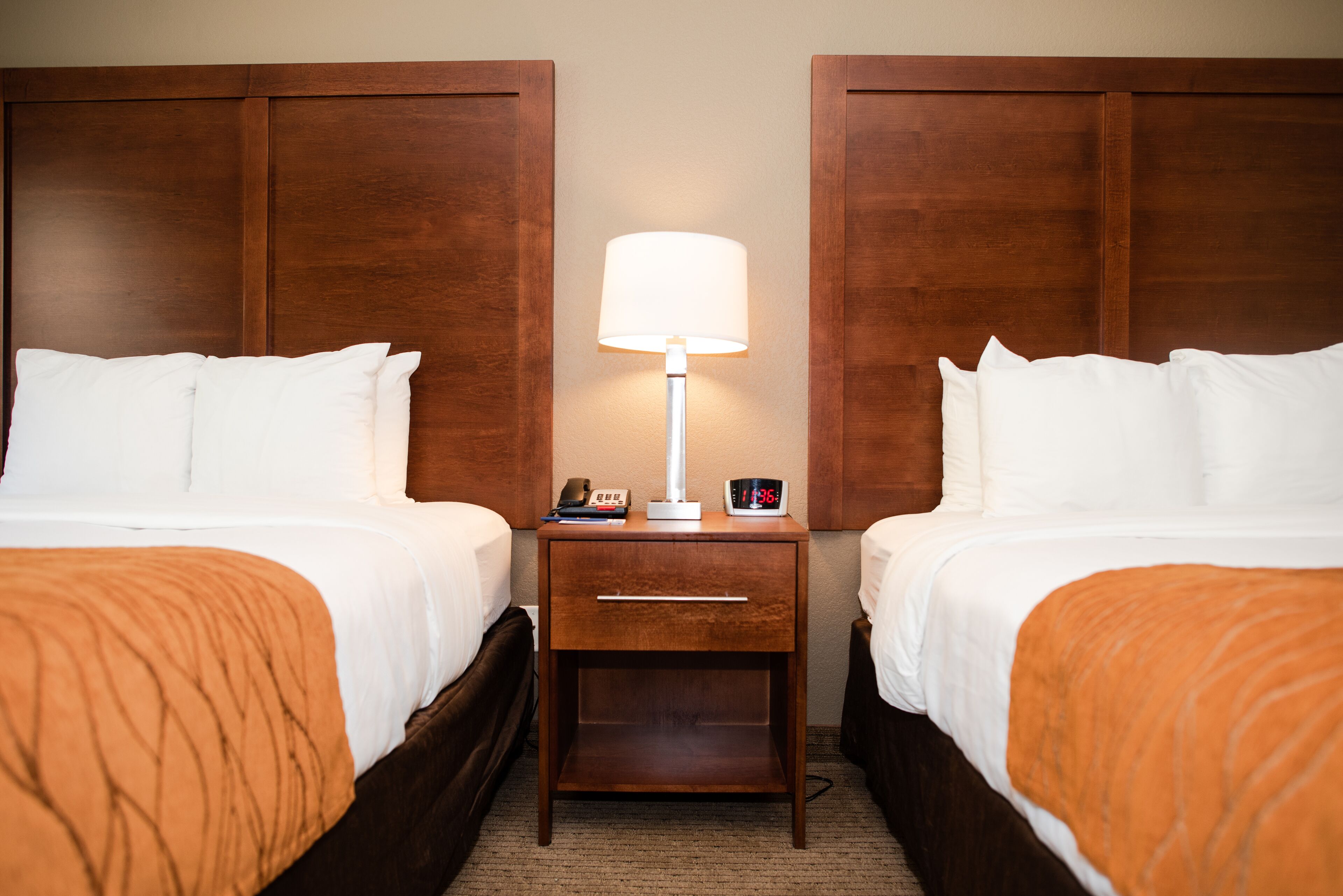A hotel room with two beds, a nightstand with a lamp, a clock, and a phone.
