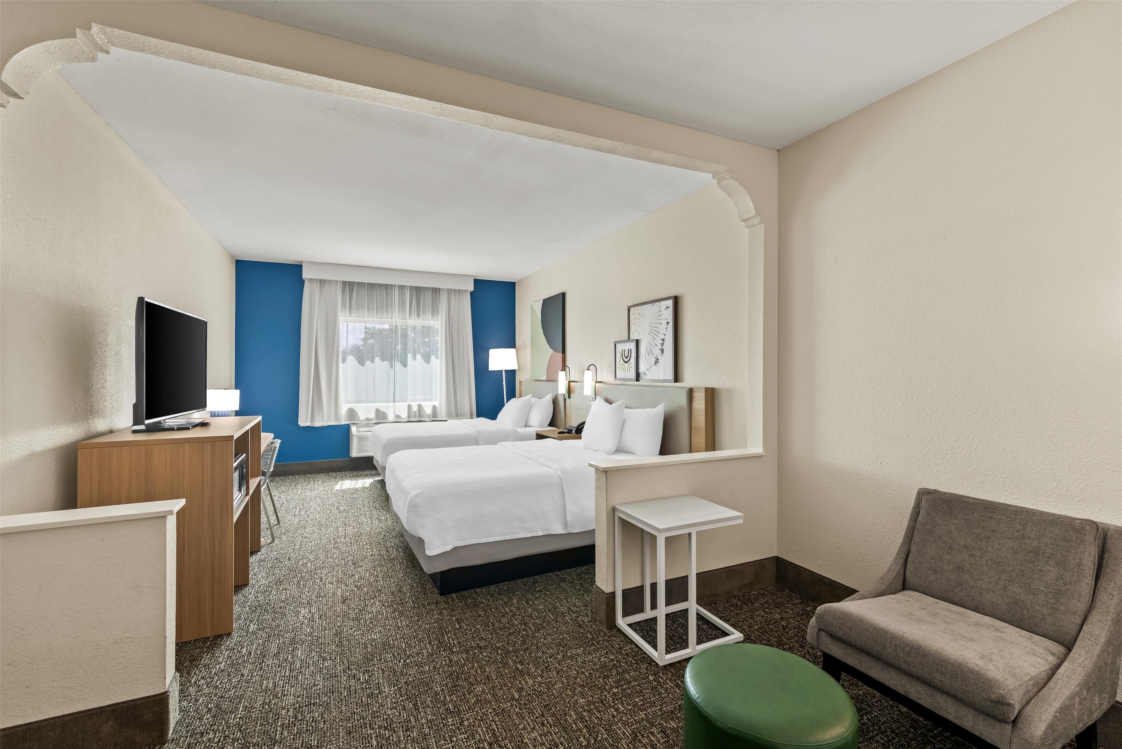 A hotel room with two beds, a desk, a television, a green ottoman, and a chair.