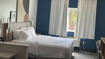 A hotel room with a bed, a desk, a chair, a window with curtains, a radiator, and a wall-mounted artwork.
