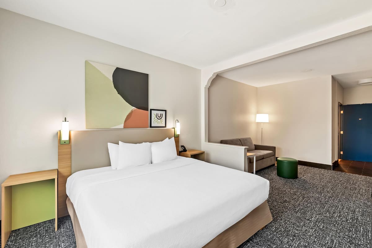 A hotel room with a bed, a desk, a chair, a green trash bin, and a door.