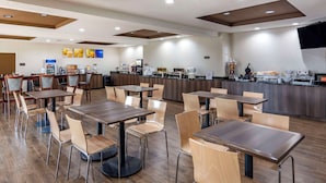 A modern restaurant with wooden tables and chairs, a counter with a beverage station, and a flat-screen TV mounted on the wall. - Comfort Suites South Park (Colonial Heights)
