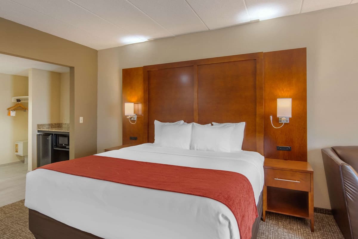 Une chambre d’hôtel avec un grand lit, une tête de lit en bois, deux lampes de chevet, une table de chevet, un bureau et, au fond, une kitchenette.