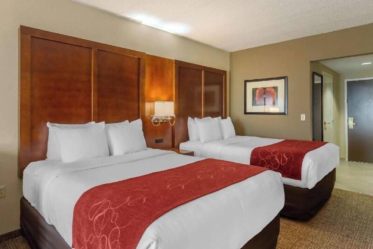 Une chambre d’hôtel avec deux lits, une tête de lit en bois, une lampe fixée au mur et un tableau encadré.