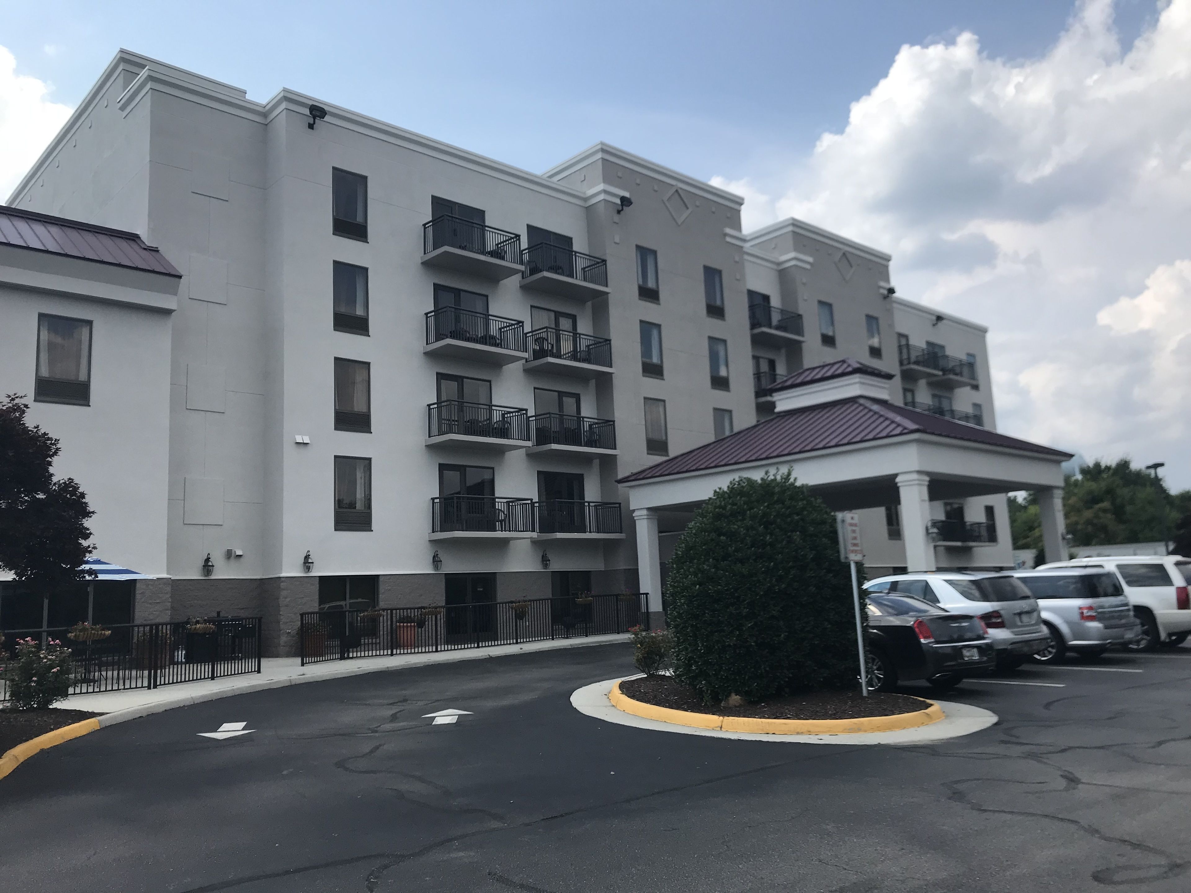 Un edificio de varios pisos con balcones, una entrada cubierta y un estacionamiento con autos.