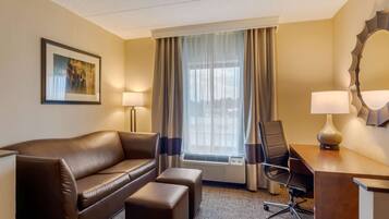 A hotel room with a brown leather sofa, a wooden desk, a chair, and a large window with curtains.