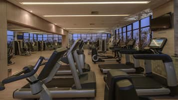 A modern gym with various cardio machines, a large window providing natural light, and a flat-screen TV mounted on the wall.