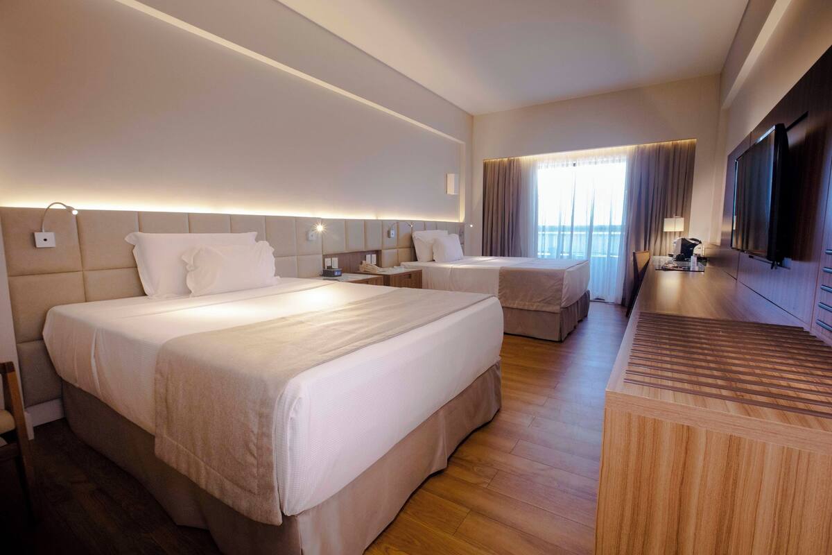 A hotel room with two beds, a wooden desk, a TV, and a large window with curtains.