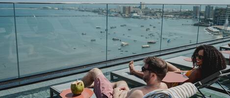 Una pareja relajándose en una terraza en la azotea con vistas a la ciudad, que incluyen un puerto deportivo con barcos.