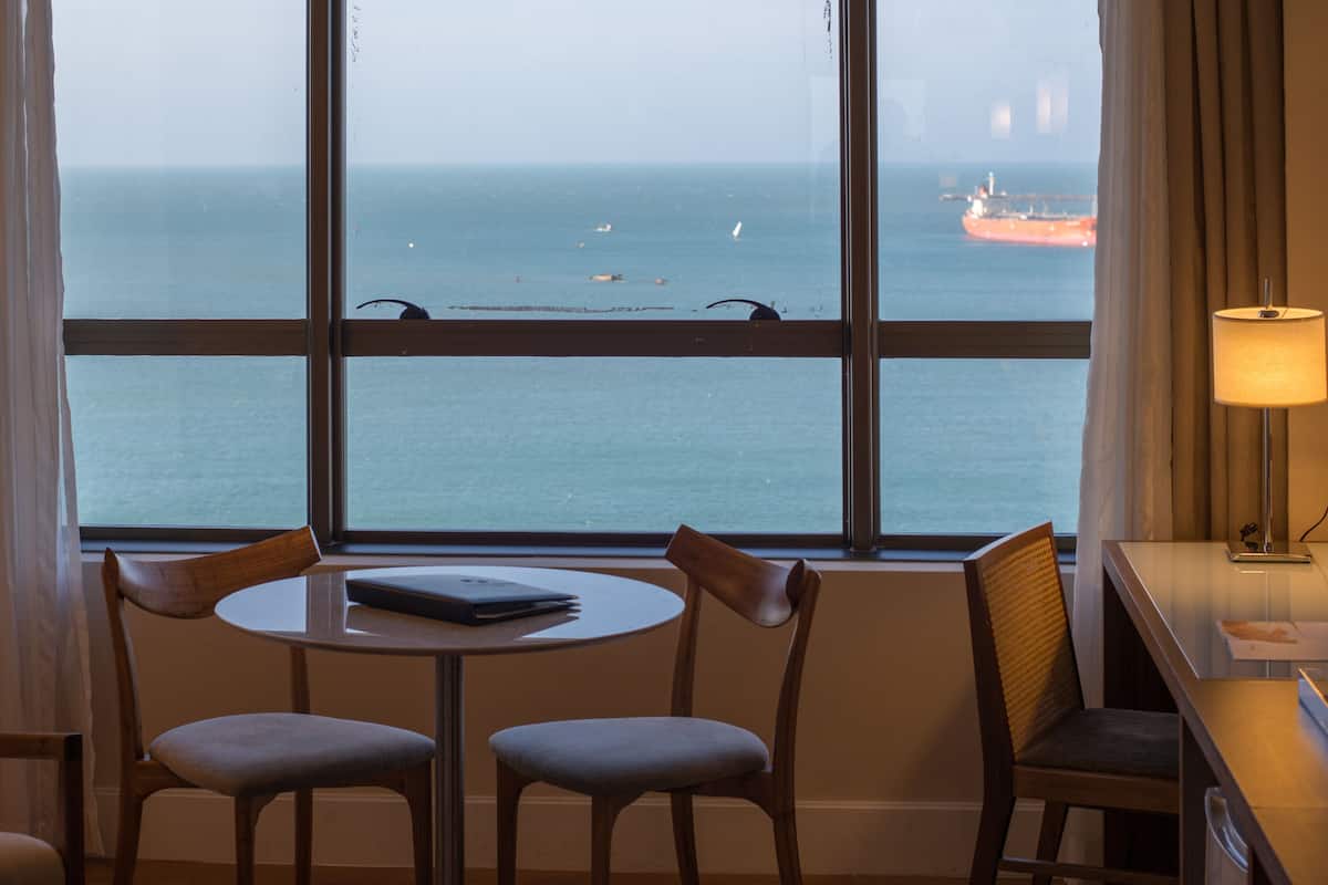 A hotel room with a large window offering a view of the sea and a ship. There is a round table with two chairs and a lamp on a bedside table.
