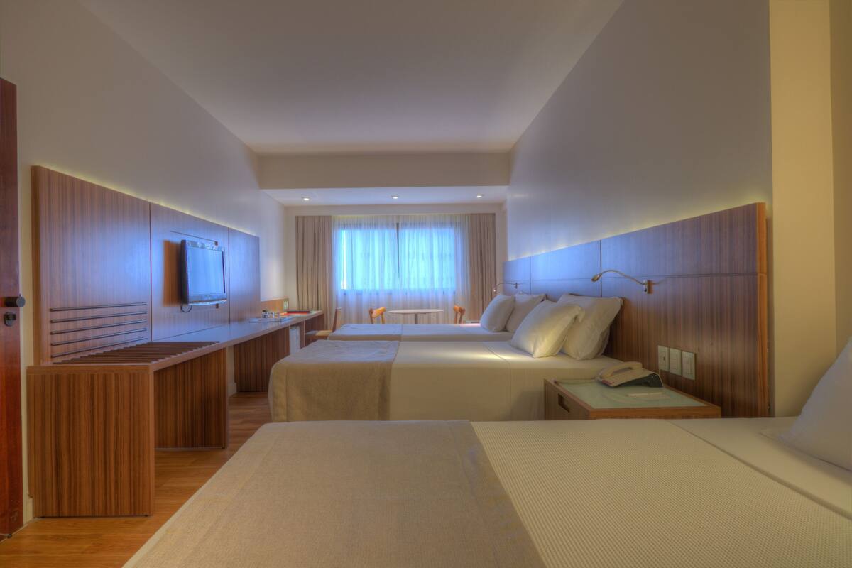 A hotel room with two beds, a desk, a TV, and a window with curtains.