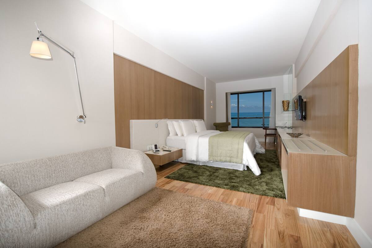 A modern hotel room with a bed, a sofa, a desk, and a view of the sea.