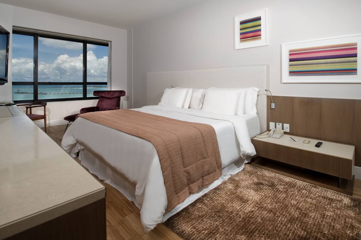 A modern hotel room with a large bed, a TV, a desk, and a view of the ocean.
