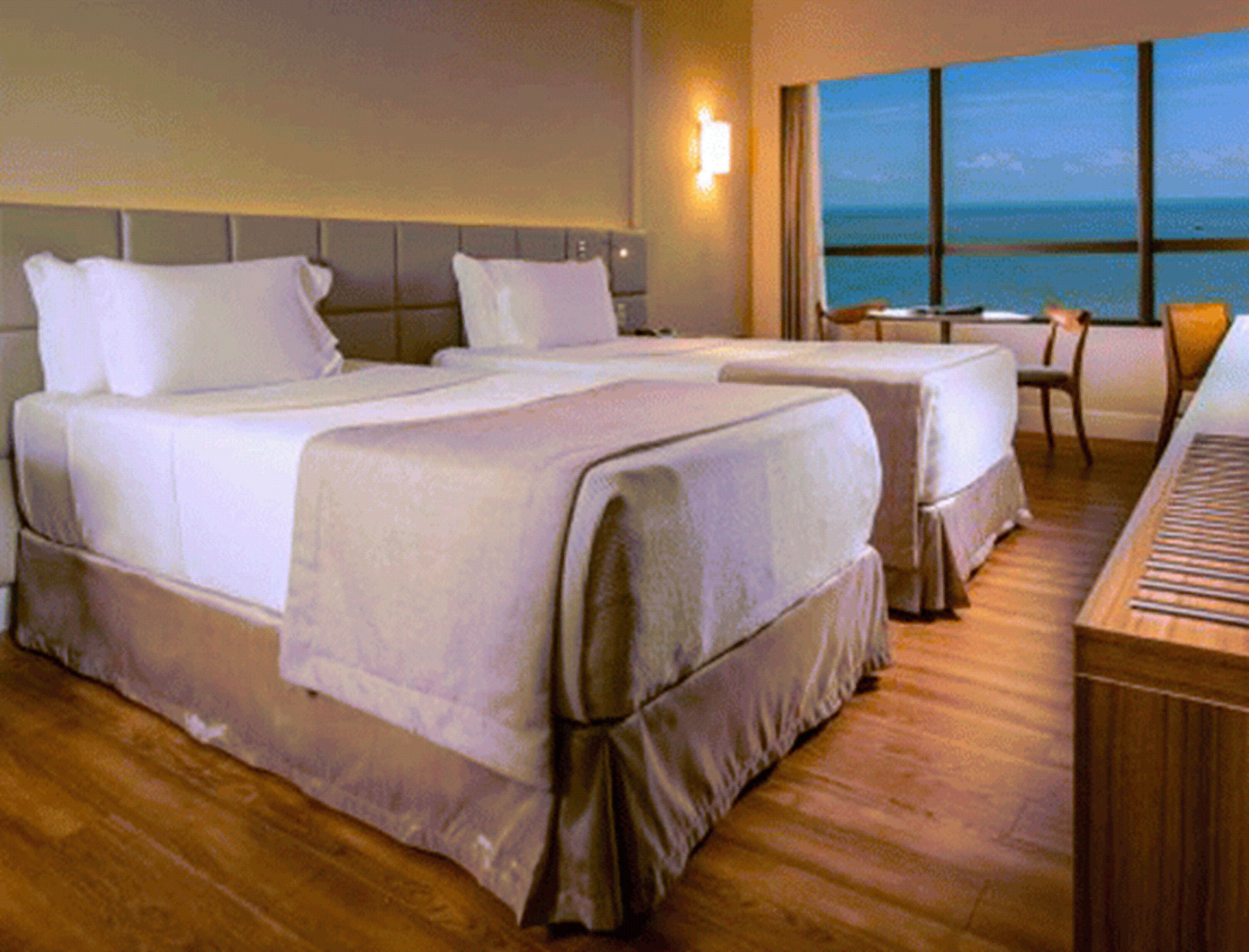 A hotel room with two beds, a piano, a small table, and a view of the ocean.