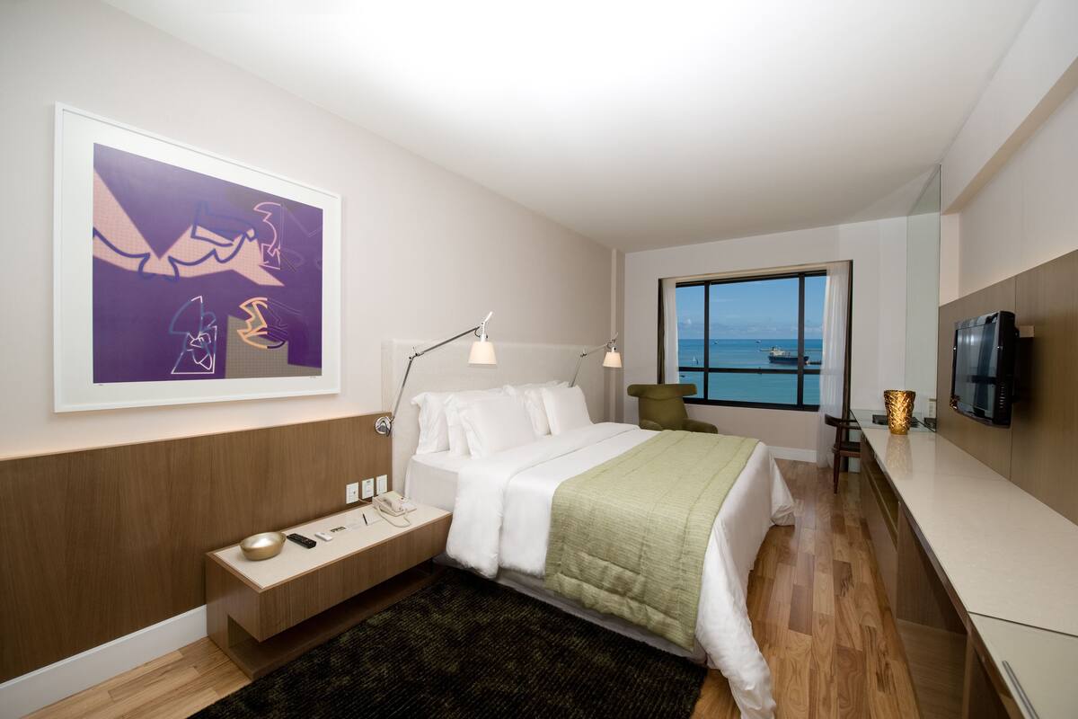 A modern hotel room with a bed, bedside table, lamp, and a large window offering a sea view.