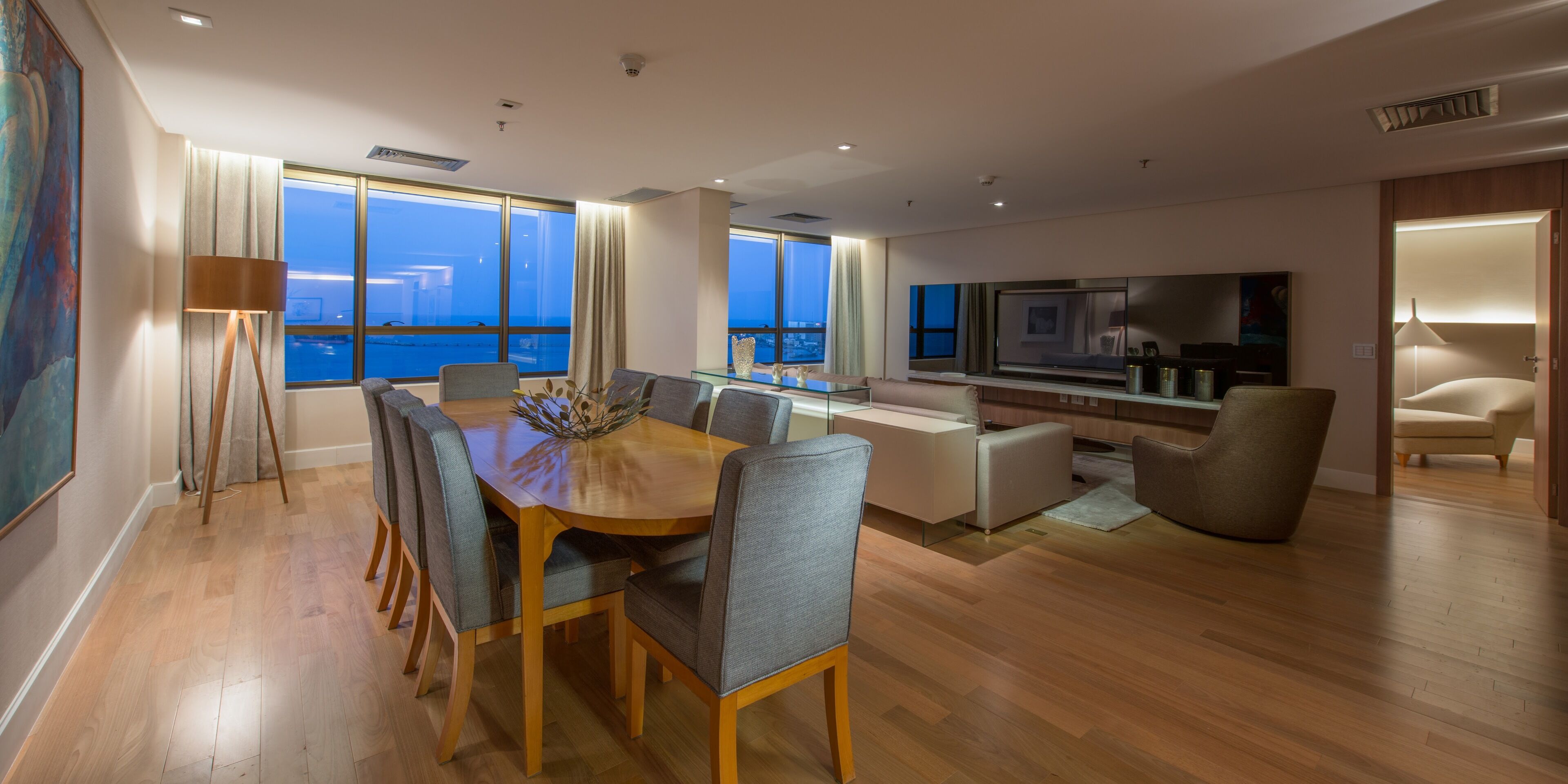 Uma sala de estar moderna com uma mesa de jantar de madeira, cadeiras estofadas cinzas, uma grande televisão de tela plana e vista para o mar através de amplas janelas.