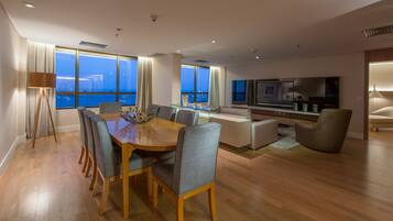 Uma sala de estar moderna com uma mesa de jantar de madeira, cadeiras estofadas cinzas, uma grande televisão de tela plana e vista para o mar através de amplas janelas.