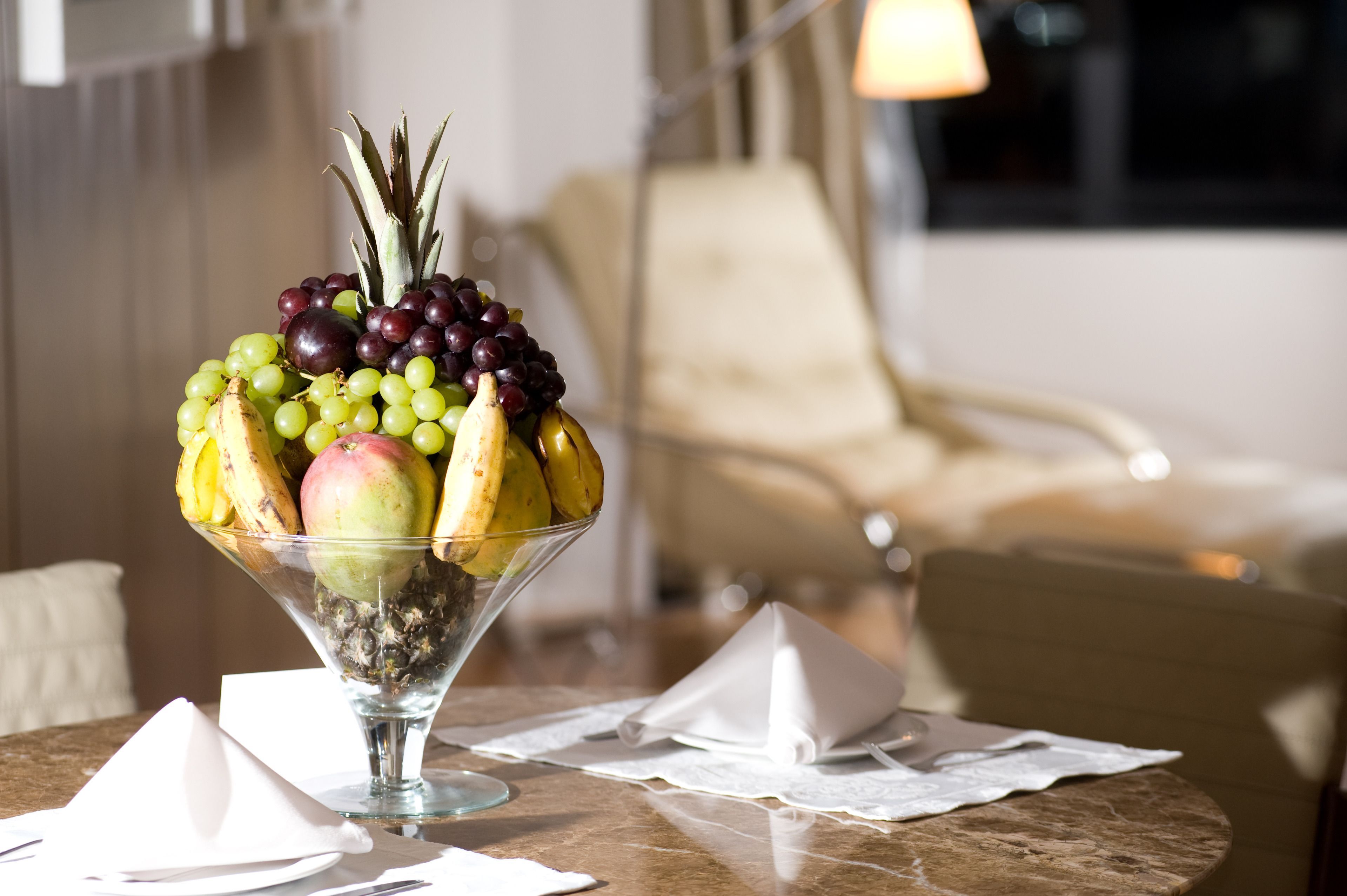Uma tigela de frutas sobre uma mesa, com uma cadeira e uma lâmpada ao fundo.